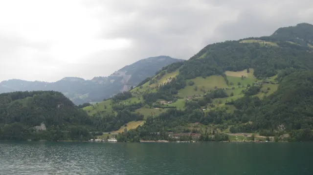 Green fields and wooded slopes rise above the lake beneath a pale, cloudy sky.