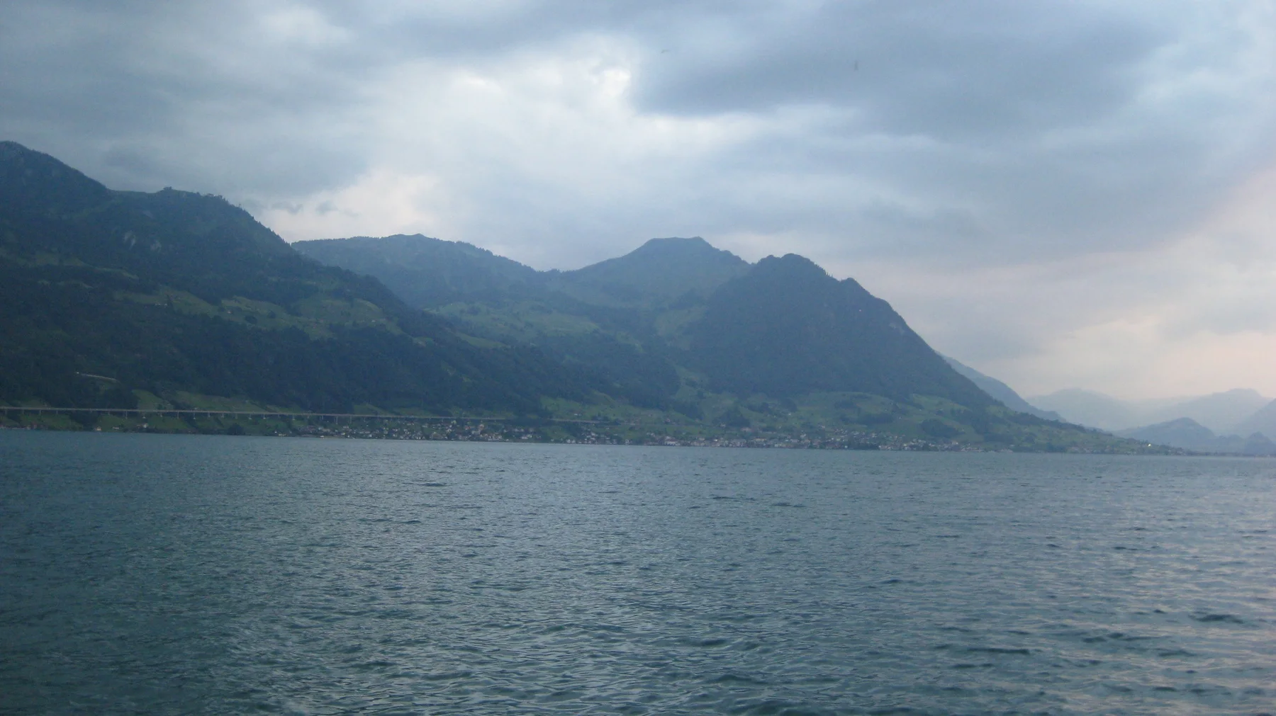 Clouds shade a long mountain shoreline above rippled lake water.