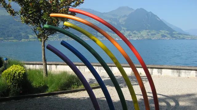 Curved rainbow-colored poles frame a lakeside promenade and mountains beyond.