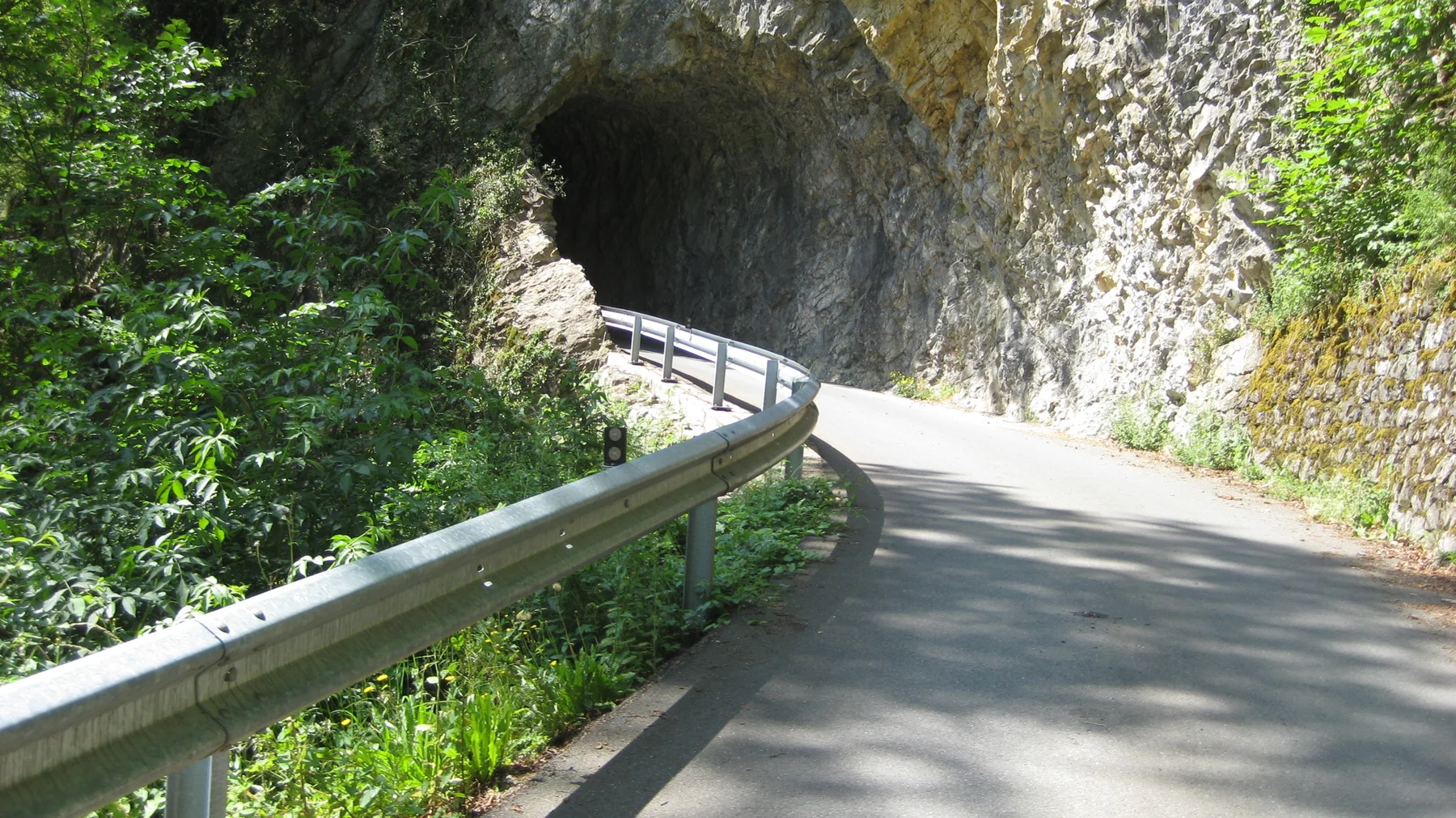 A narrow paved road curves past a guardrail into a tunnel through the rock.