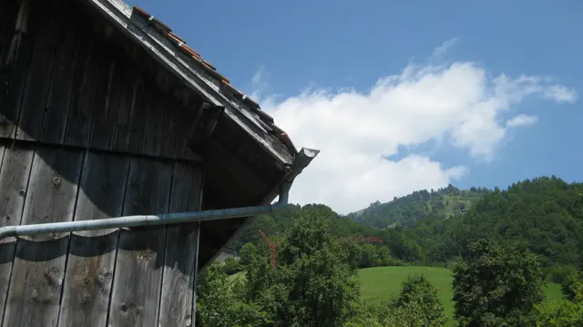 The weathered wall and roof of a wooden shed frame a green hillside and sky.