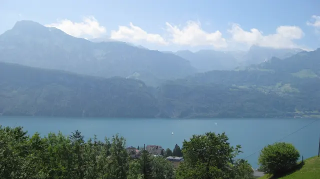 Lakeside buildings and trees lie below a blue lake and hazy mountain peaks.