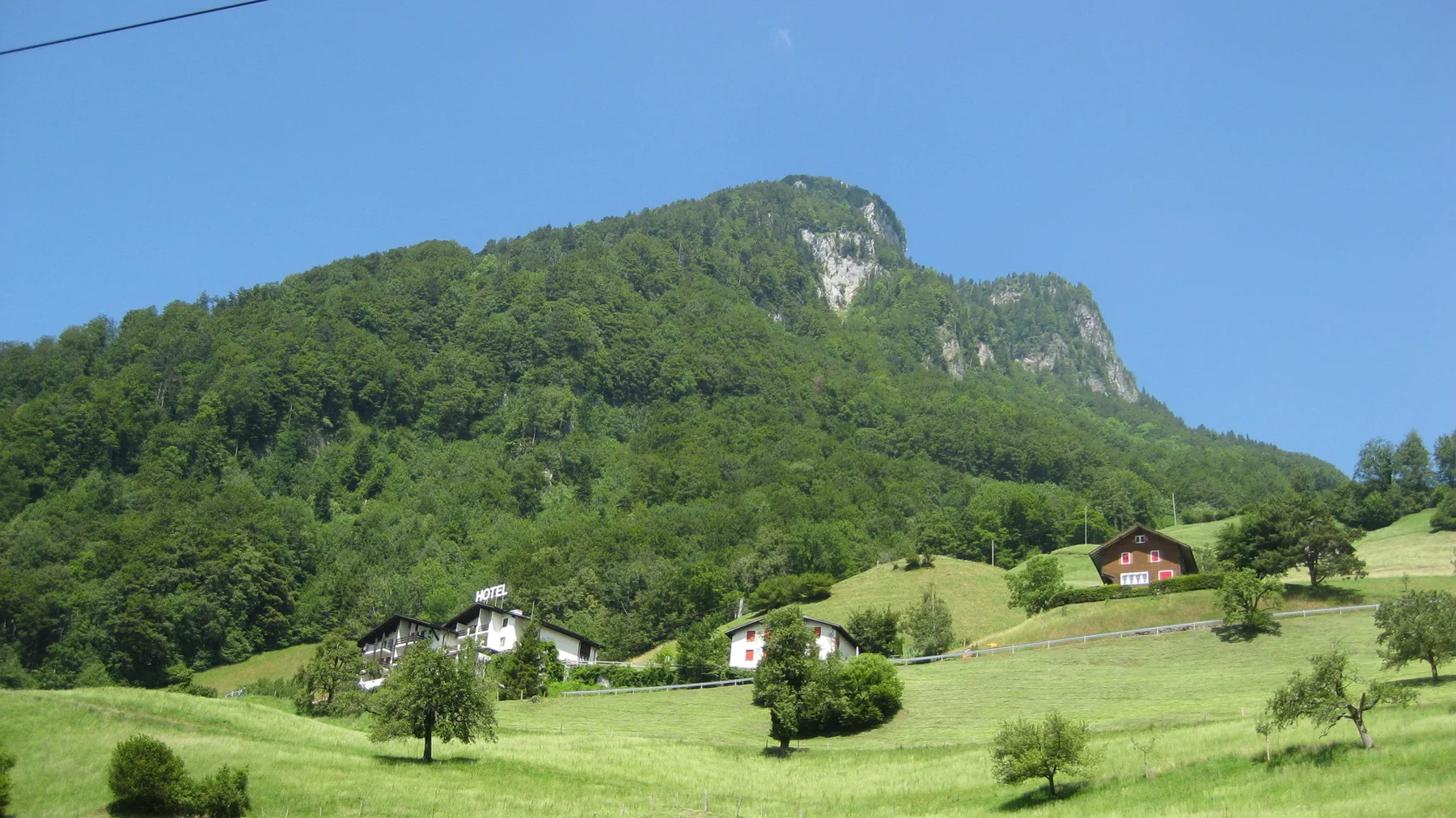 Houses and orchard trees dot green fields below a forested, rocky ridge.