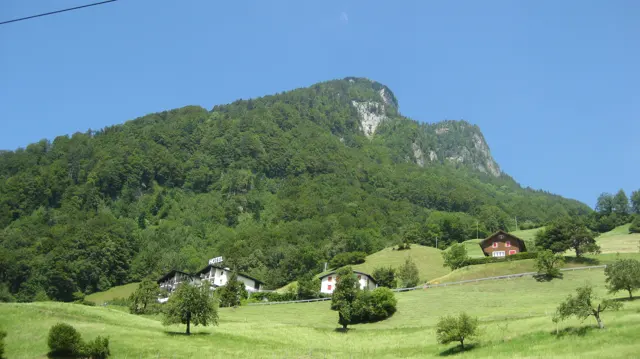 Houses and orchard trees dot green fields below a forested, rocky ridge.