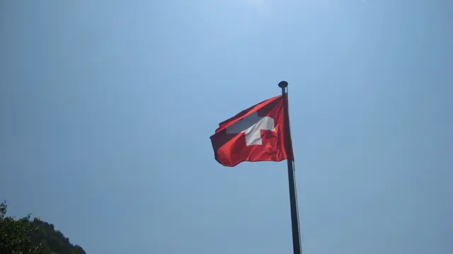 A red Swiss flag flies from a tall pole against a clear blue sky.