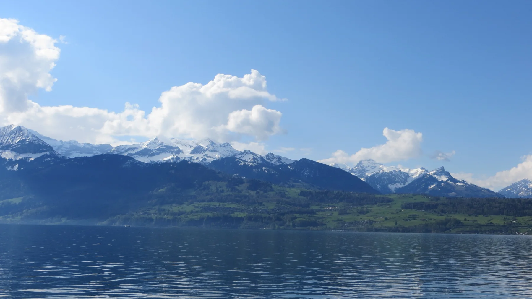 A snow-lined mountain range stretches across a rippled blue lake beneath white clouds.