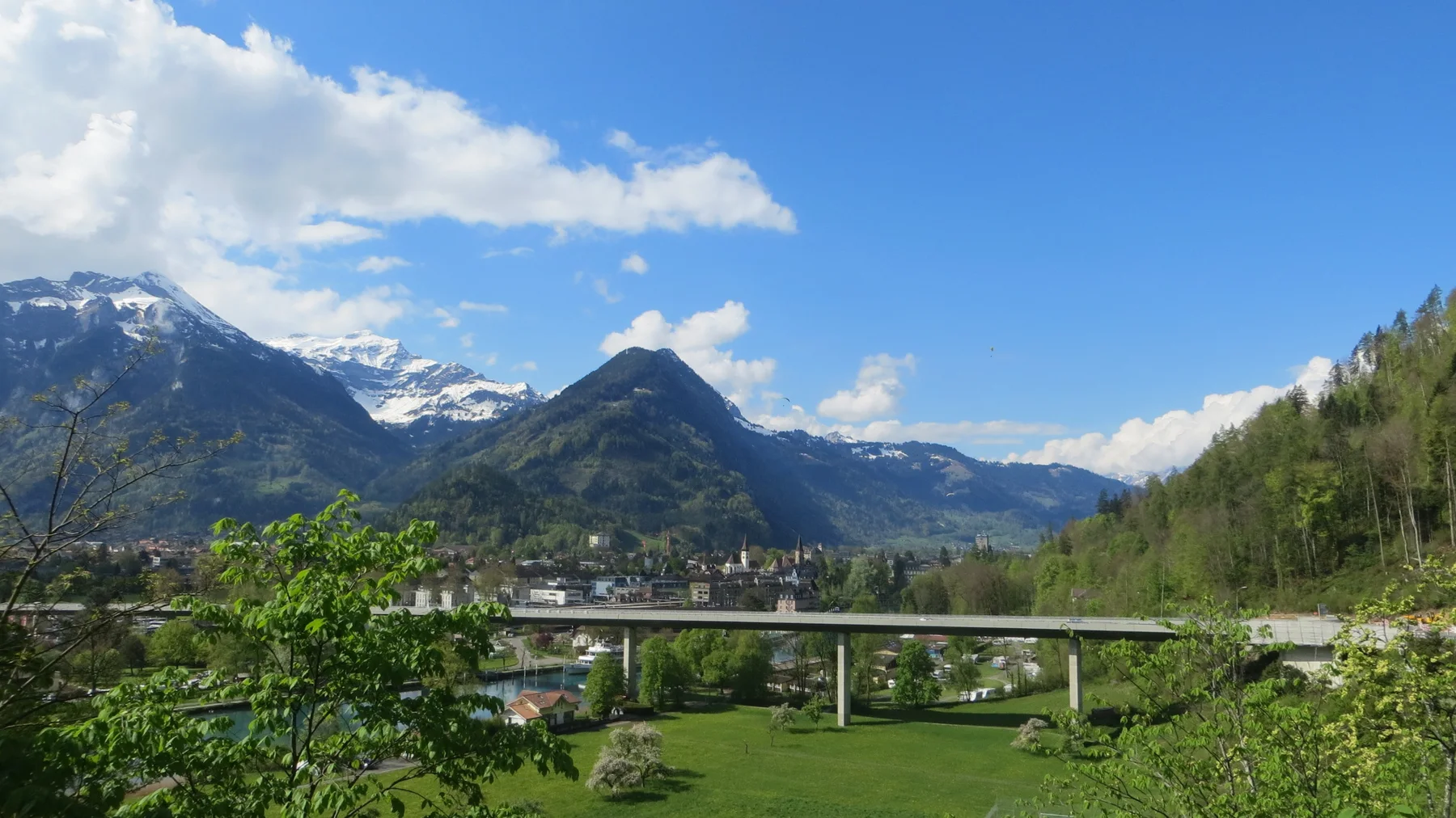 A long bridge crosses a green valley below pointed mountains and snowy ridges.