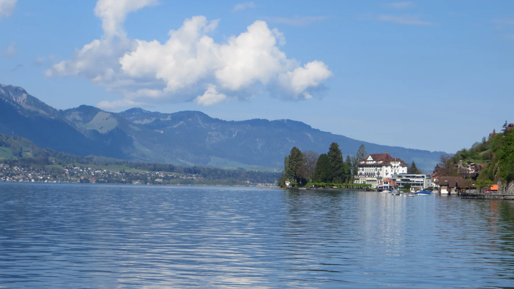 White lakeside buildings and trees stand on a point beneath distant mountains.