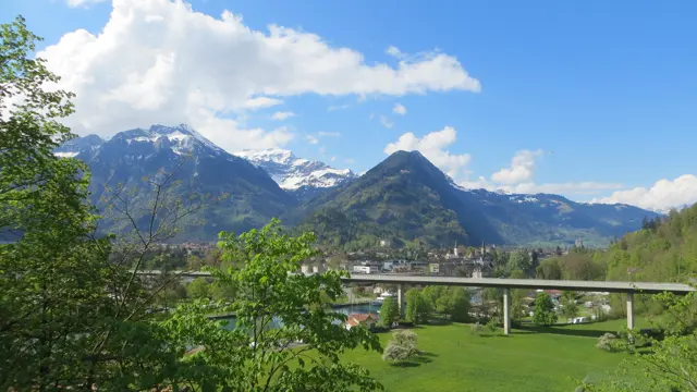 A road bridge spans green fields with a mountain settlement and snowy peaks beyond.