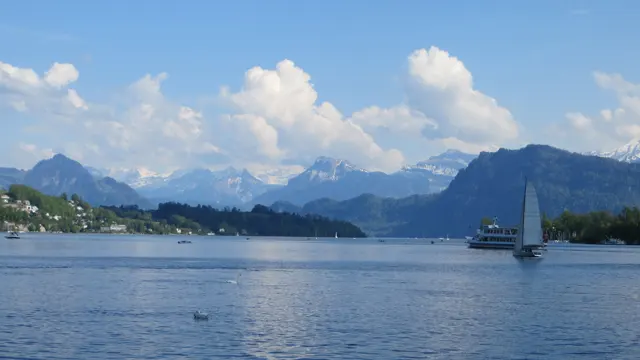 A sailboat moves across blue water with shoreline buildings and mountain peaks behind it.