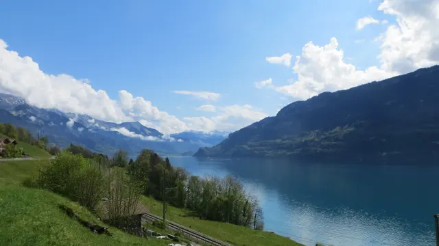 A railway follows a grassy slope above still water and a distant mountain valley.