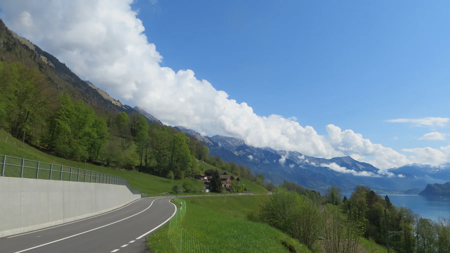 A paved road curves beside a concrete wall above a green slope and blue lake.