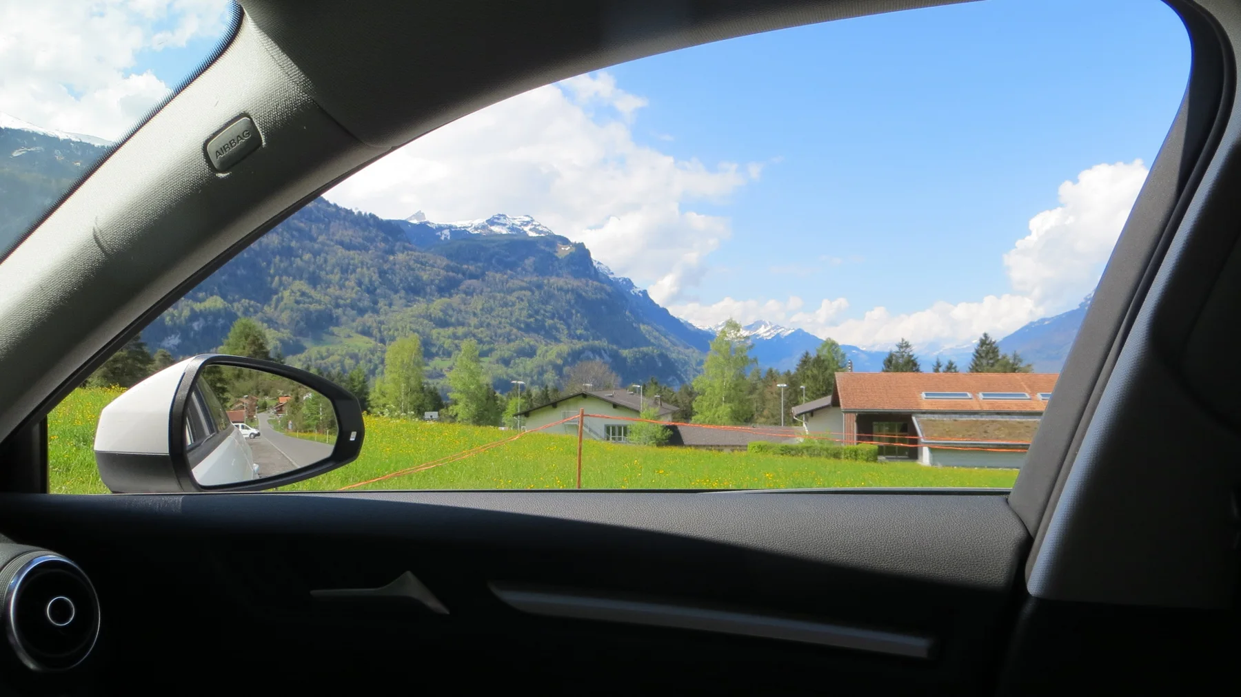 A car's passenger window frames green fields, a wooden building and snowy mountains.