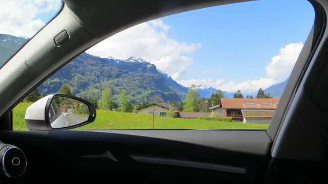 A car's passenger window frames green fields, a wooden building and snowy mountains.