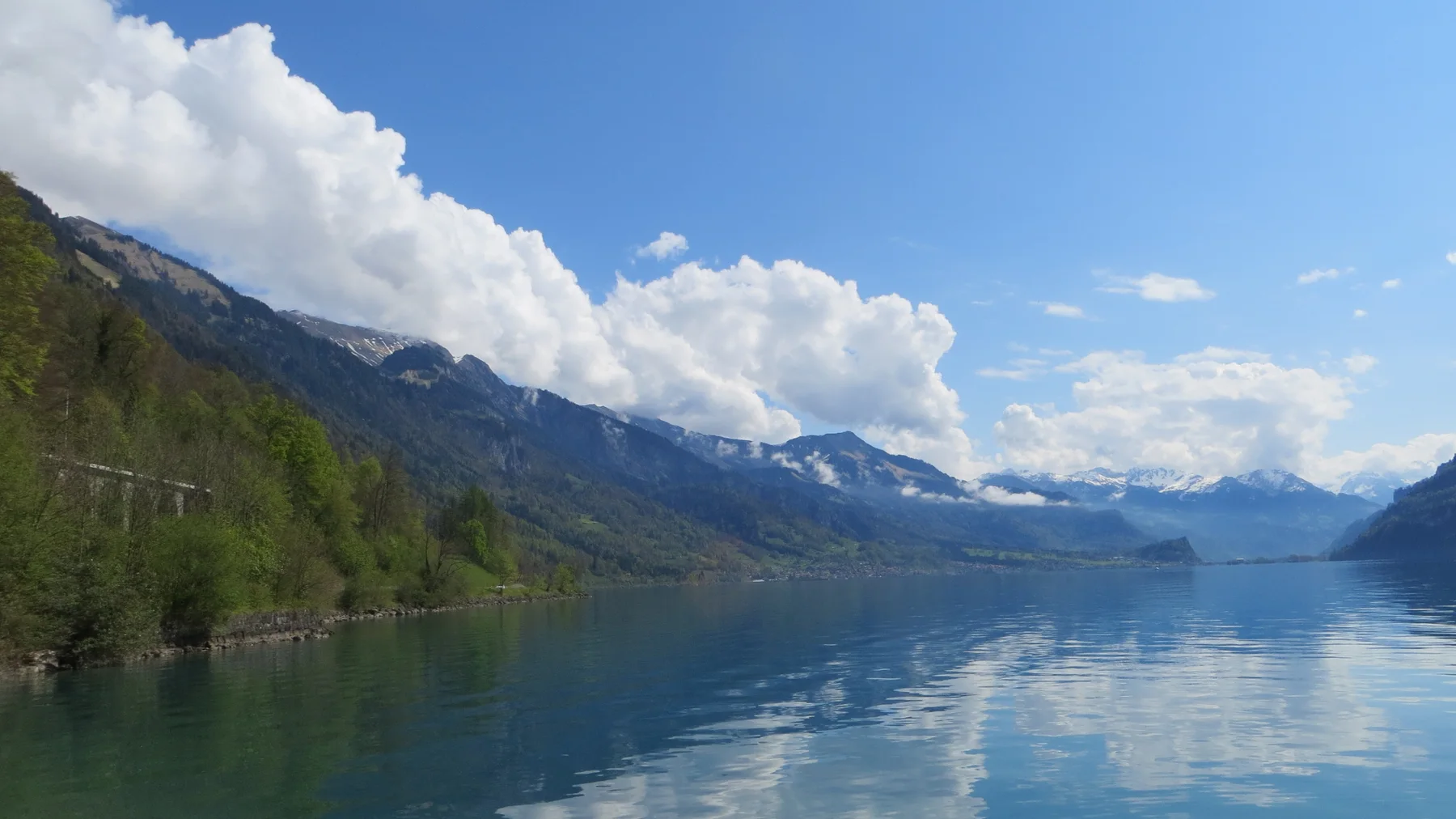 A green mountain shore leads toward snowy peaks reflected in the lake.