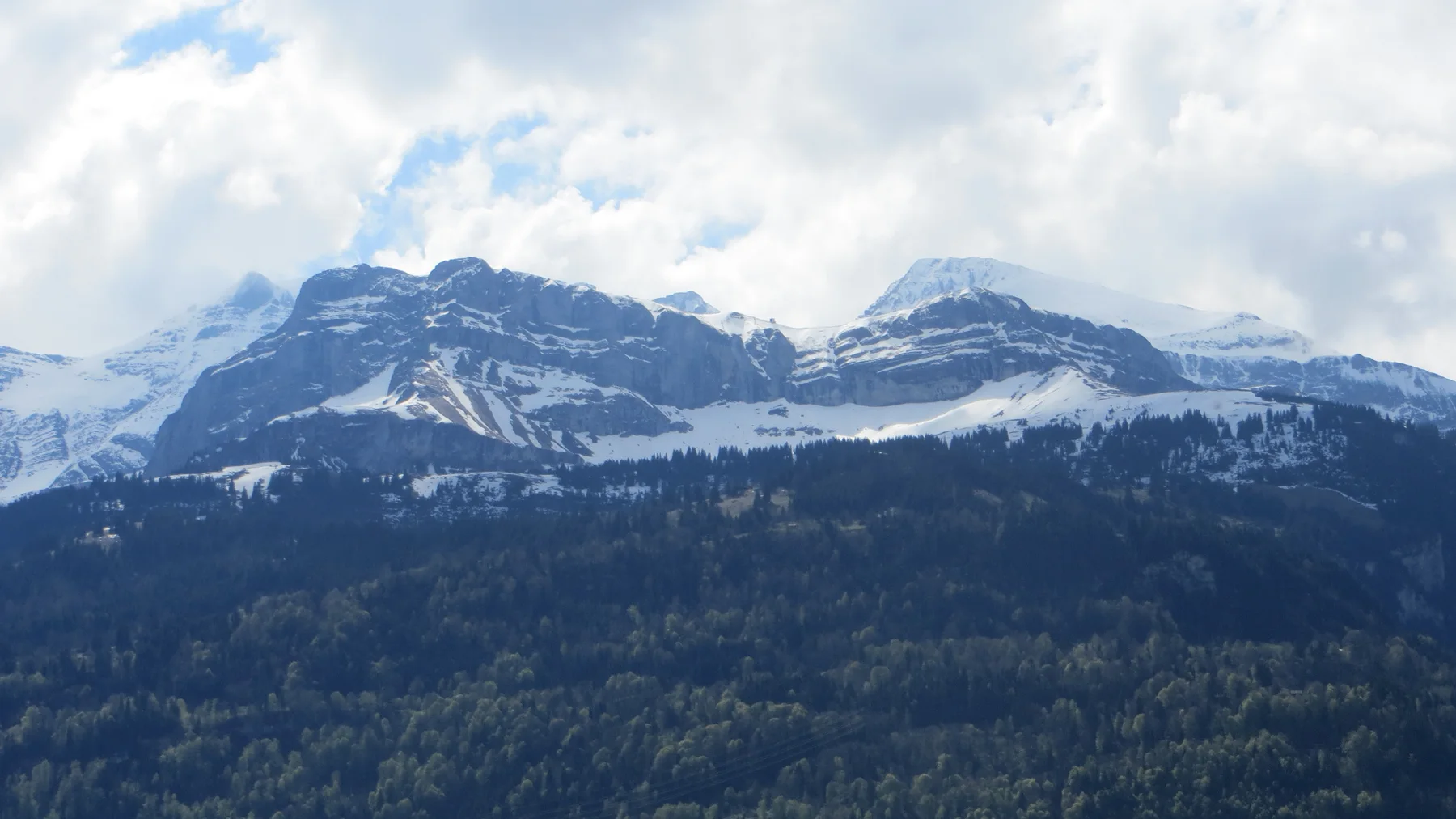 Snow traces the ledges of layered mountain peaks above a dark forest.