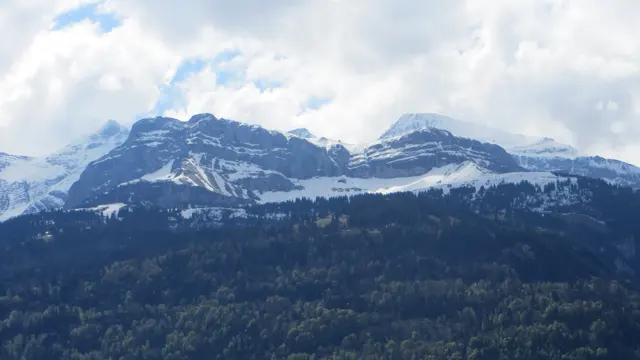 Snow traces the ledges of layered mountain peaks above a dark forest.