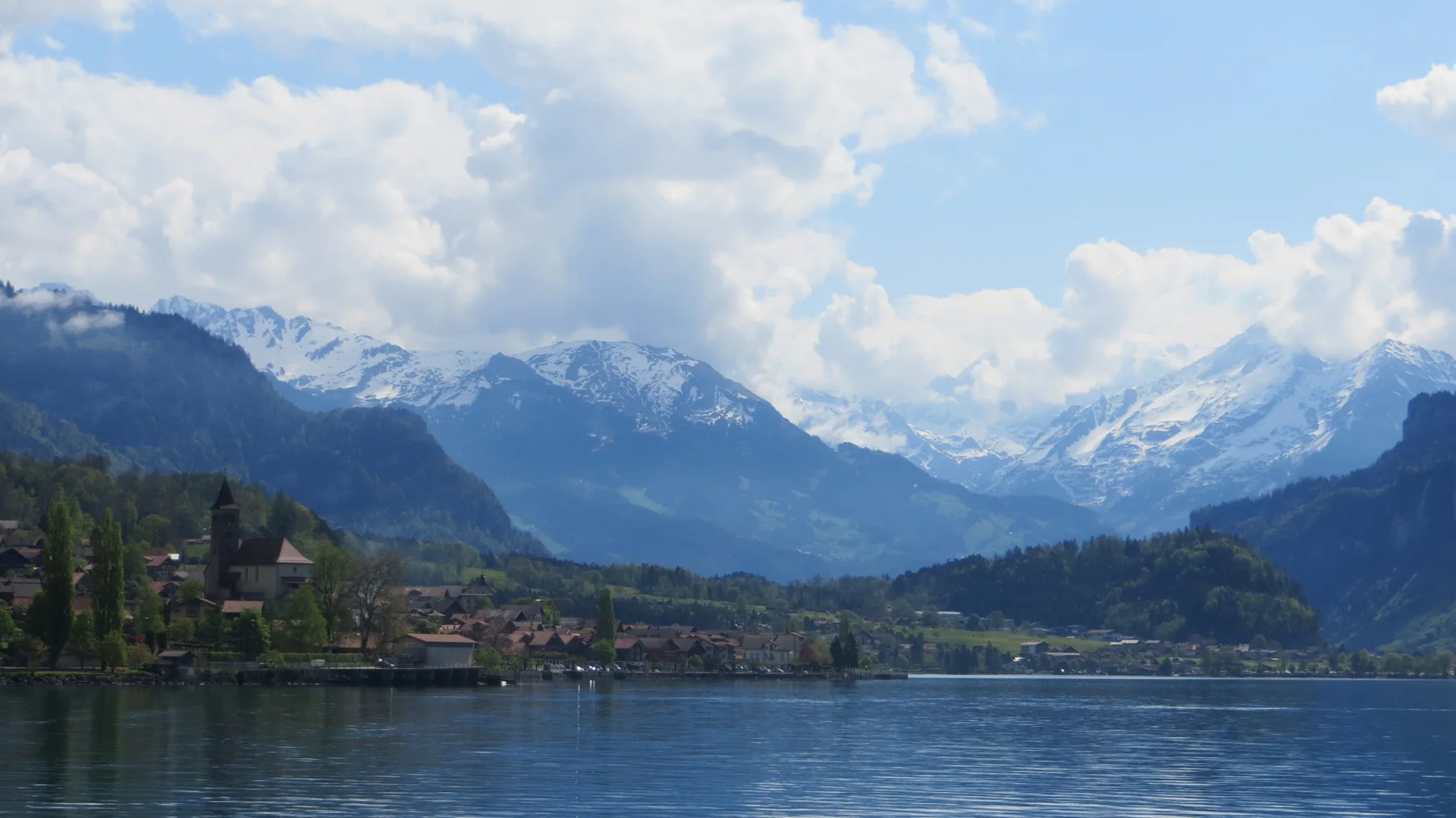 A lakeside village lies beneath a mountain valley filled with cloud and distant snow.
