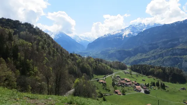 A small group of buildings sits in a green valley between woodland and snowy mountains.