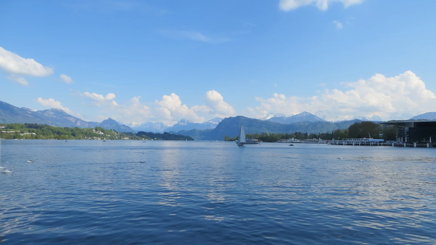 A distant white sail crosses a broad lake beneath scattered clouds and mountain peaks.