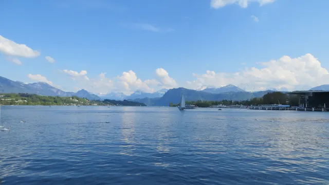 A distant white sail crosses a broad lake beneath scattered clouds and mountain peaks.