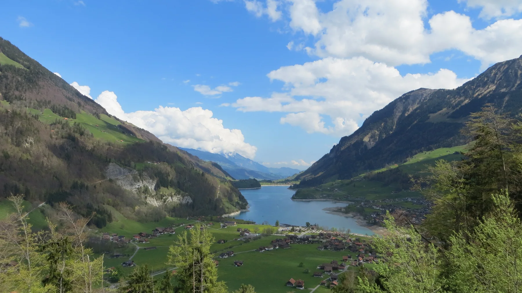 An elevated view follows green fields and a village toward a lake between steep mountains.