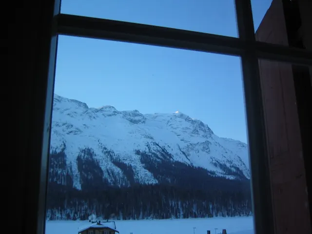 A dark window frame encloses a view of snowy mountains and a winter valley.