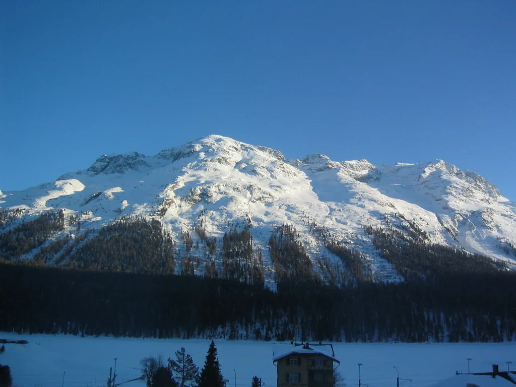 Sunlit snowy peaks rise above a dark forest and snow-covered valley.