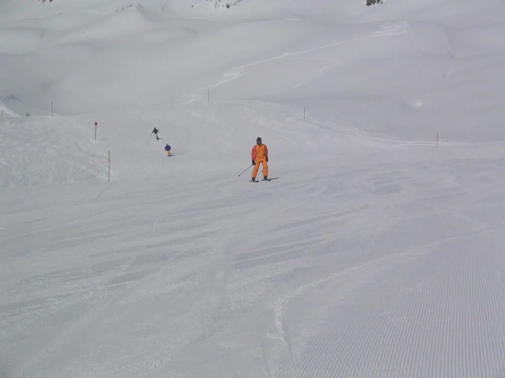A figure in orange stands on a snowy slope among faint tracks and distant skiers.