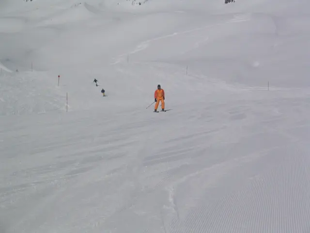 A figure in orange stands on a snowy slope among faint tracks and distant skiers.