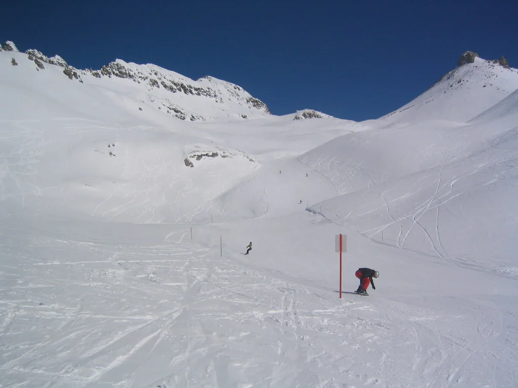 Skiers descend a broad white slope beneath a rocky mountain saddle.