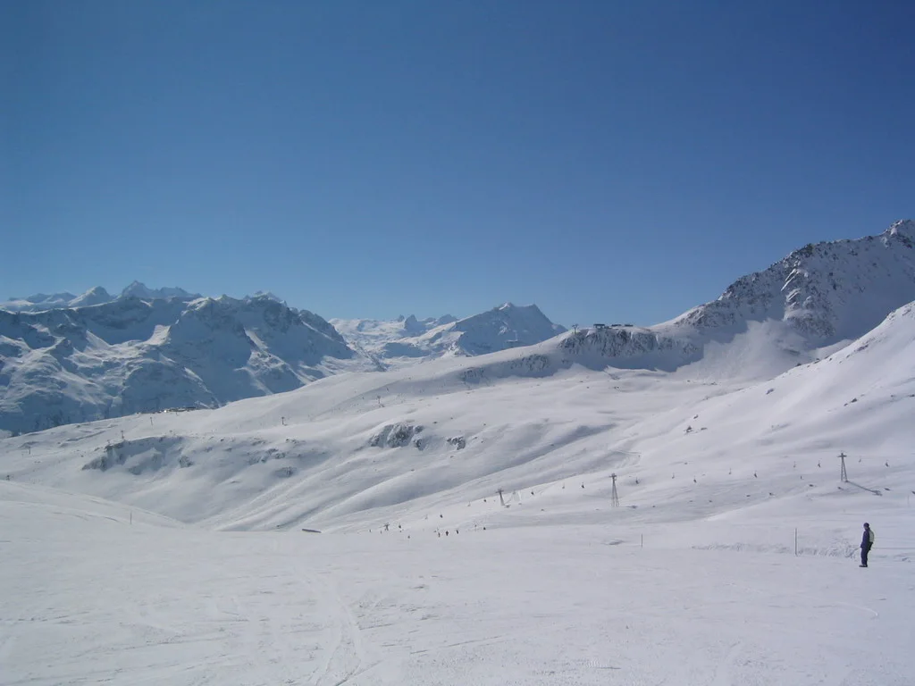 A snowy basin opens toward a range of jagged mountains under clear blue sky.
