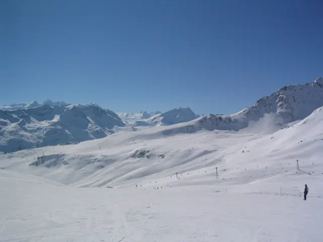 A snowy basin opens toward a range of jagged mountains under clear blue sky.