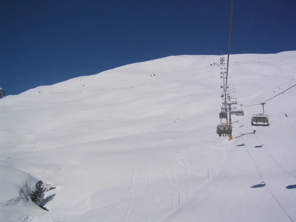 Chairlift seats ascend beside a tower over a smooth snowy hillside.