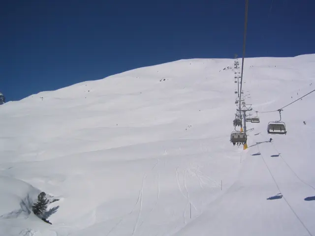 Chairlift seats ascend beside a tower over a smooth snowy hillside.