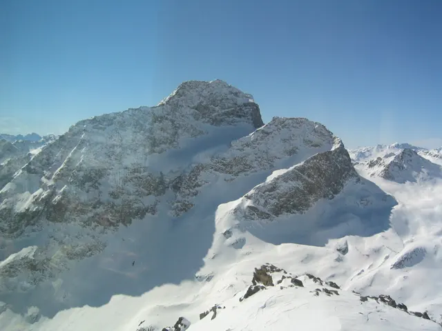 A rugged rock summit rises above deep snow and distant mountain peaks.