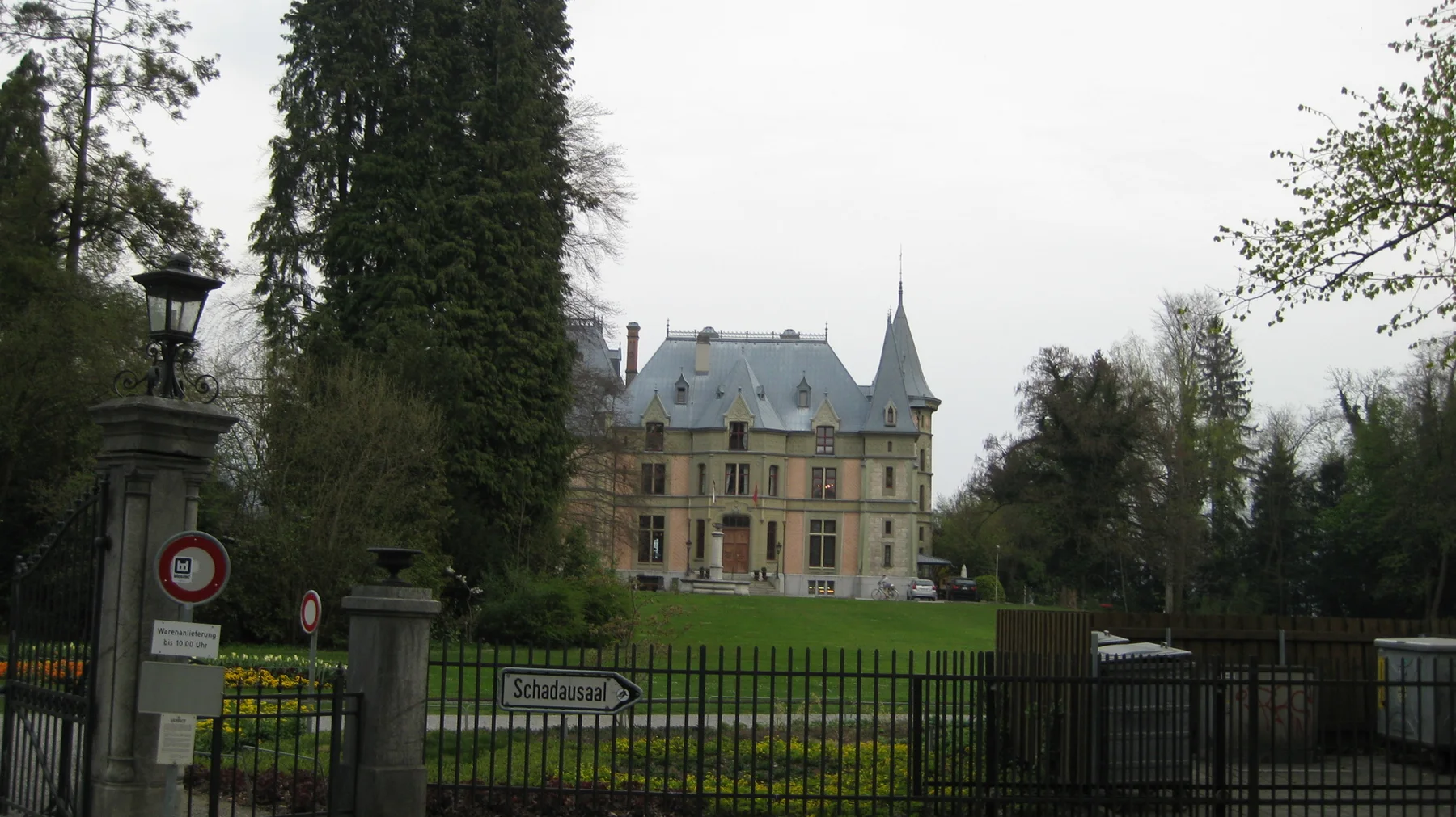 An ornate house with steep roofs stands beyond iron gates and a green lawn.