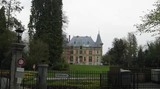 An ornate house with steep roofs stands beyond iron gates and a green lawn.