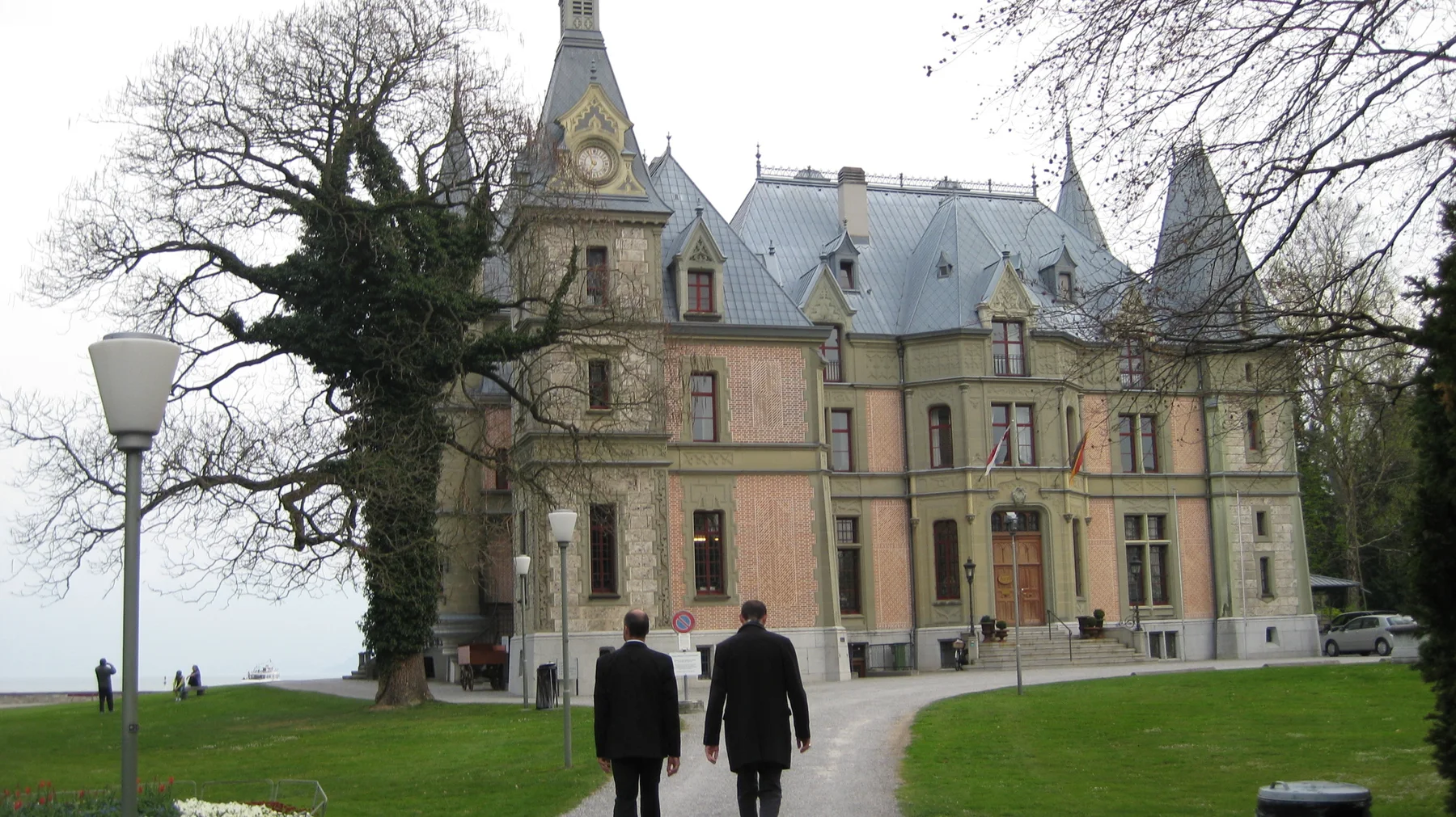 Two people walk toward an ornate lakeside building with steep roofs and pointed turrets.