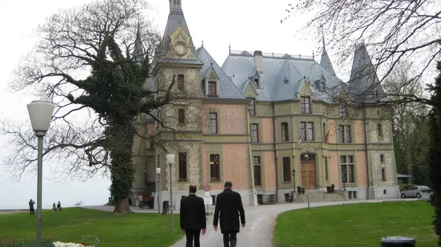Two people walk toward an ornate lakeside building with steep roofs and pointed turrets.