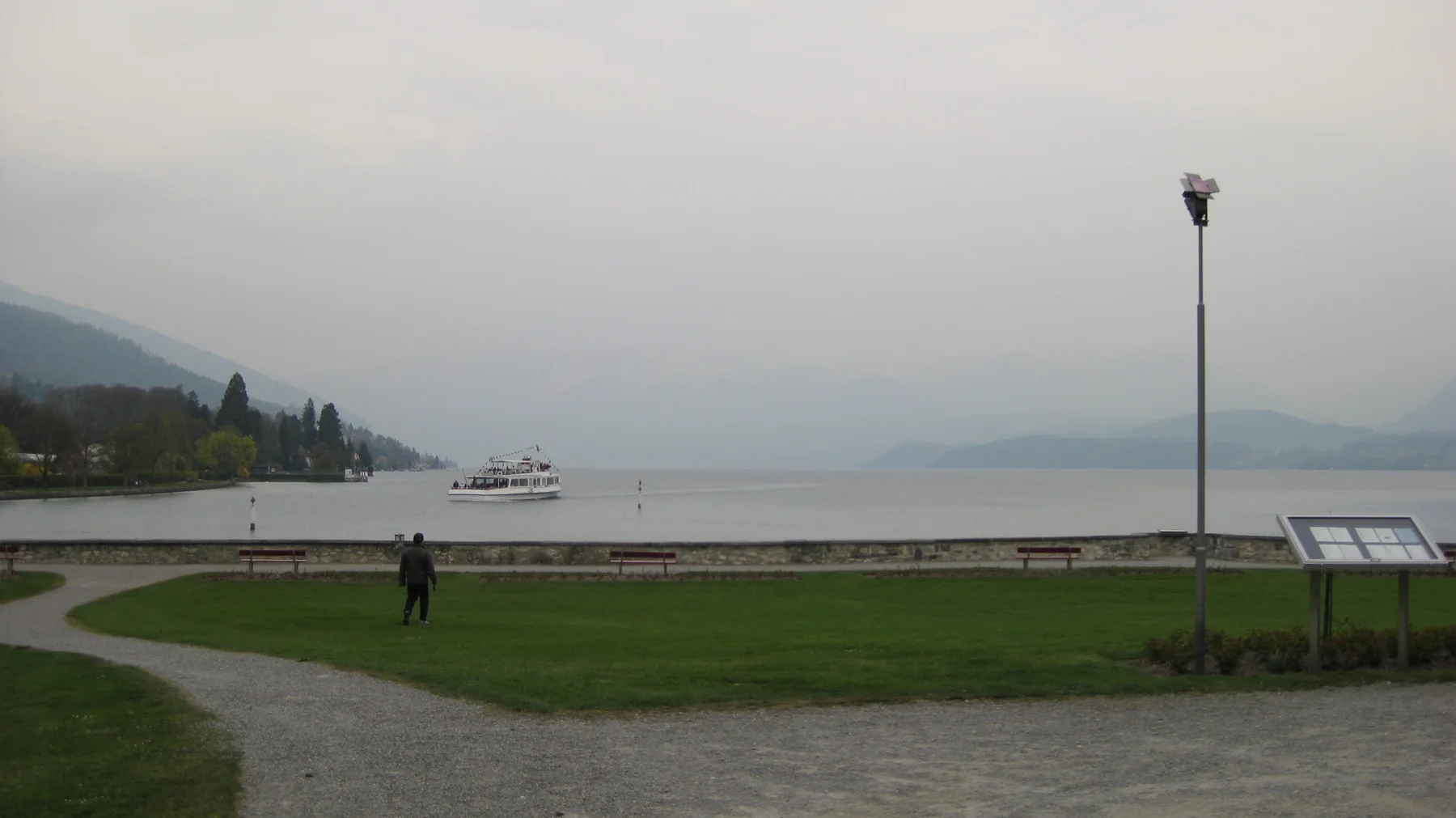 A curved gravel path and green lawn meet a quiet lake beneath hazy mountains.
