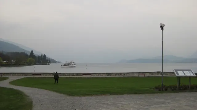 A curved gravel path and green lawn meet a quiet lake beneath hazy mountains.