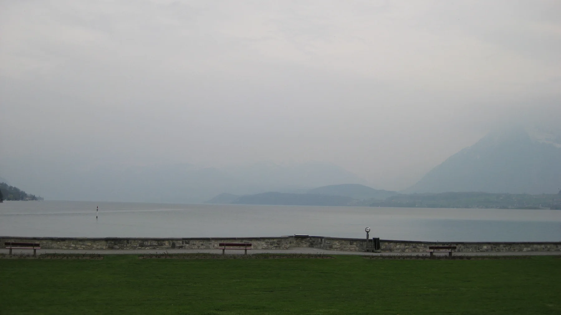 A low stone shore and green lawn face a lake fading into mountain haze.