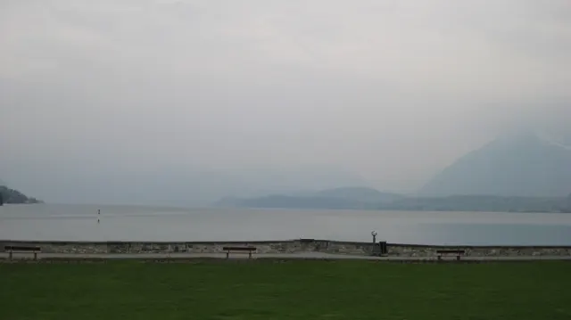 A low stone shore and green lawn face a lake fading into mountain haze.