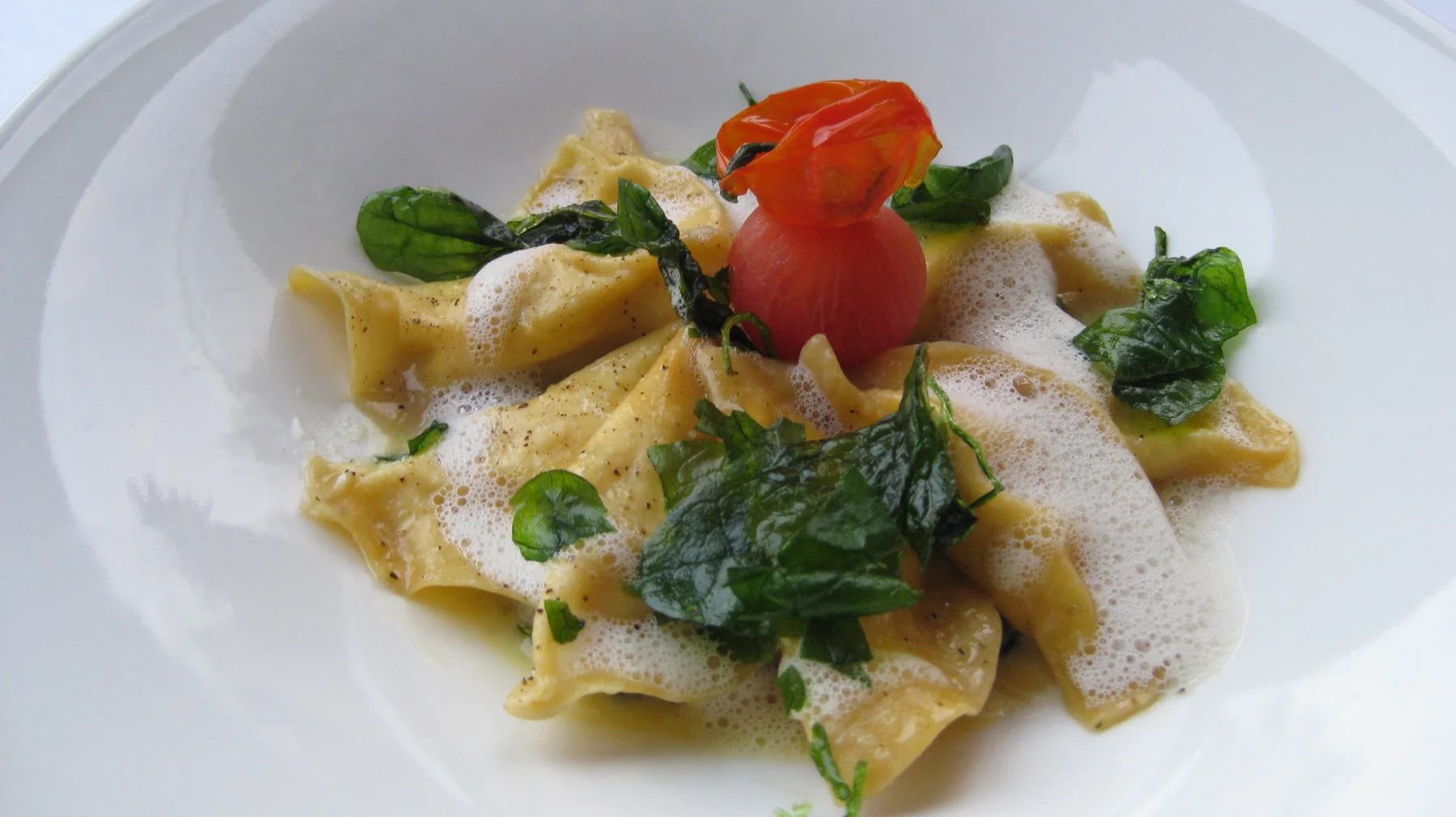Folded pasta, green vegetables and a red tomato sit in a wide white bowl.