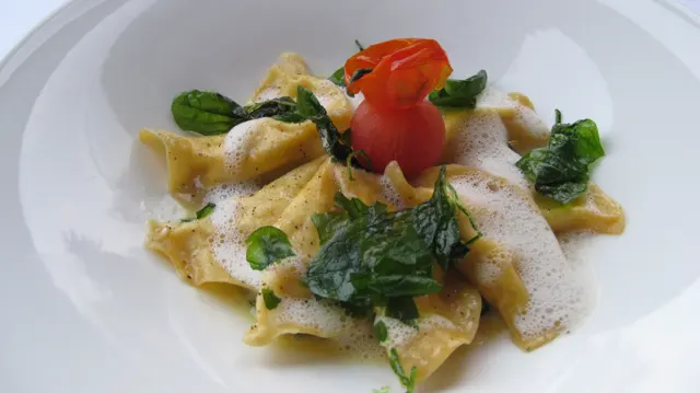 Folded pasta, green vegetables and a red tomato sit in a wide white bowl.