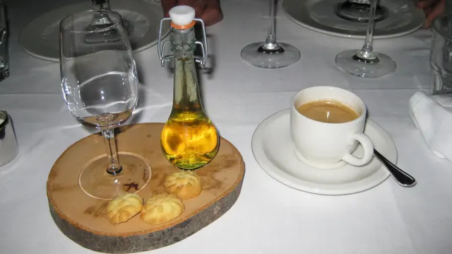 A white coffee cup stands beside glasses and a small bottle of golden liquid on a wooden board.