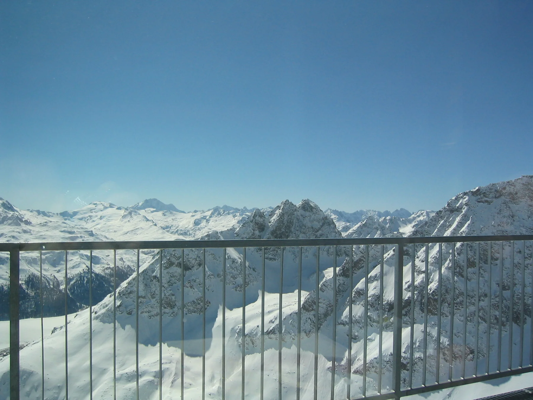 A metal balcony railing overlooks a wide range of snowy mountains under blue sky.