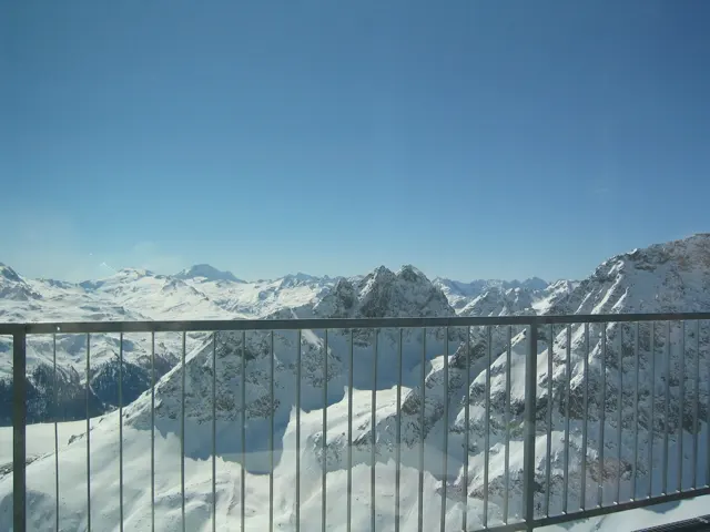 A metal balcony railing overlooks a wide range of snowy mountains under blue sky.