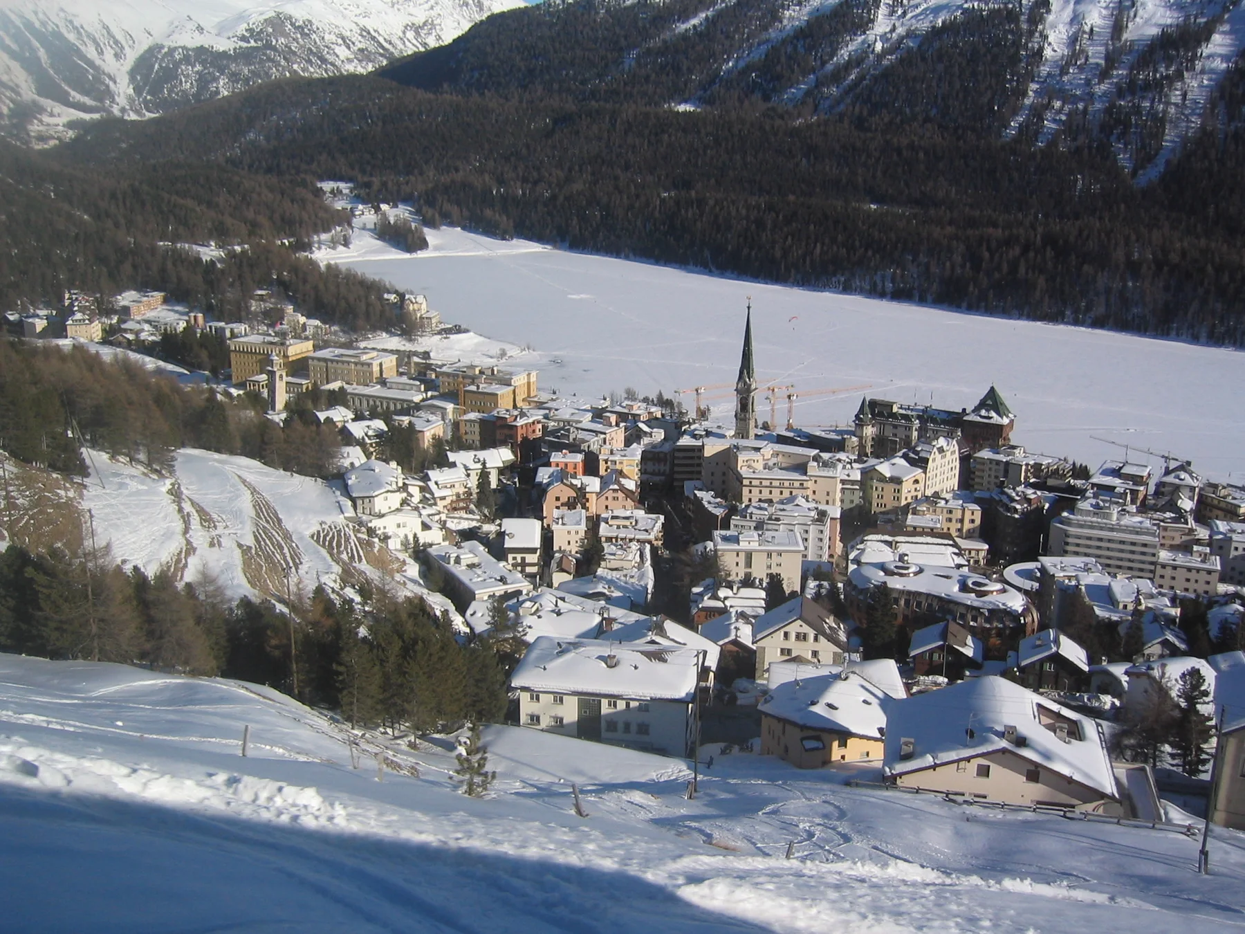 Snow-covered town roofs and a church spire lie beside a pale frozen lake.
